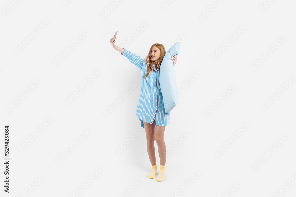 Happy young girl in pajamas holds a soft pillow and smiles while taking a selfie on a white background Happy young girl in pajamas holds a soft pillow and smiles while taking a selfie on a white background