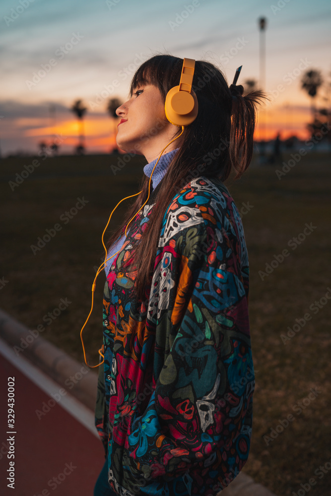 Young millennial melancholy woman listening to music with yellow headphone in an urban park during colorful sunset