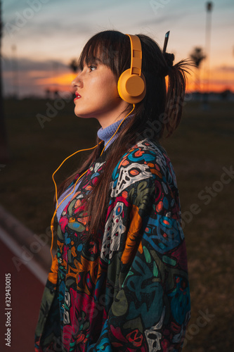 Young millennial melancholy woman listening to music with yellow headphone in an urban park during colorful sunset © ManuPadilla