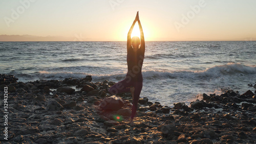 Flexible caucasian girl doing tree yoga asana at seashore at sunset. © Alexander Belinskiy