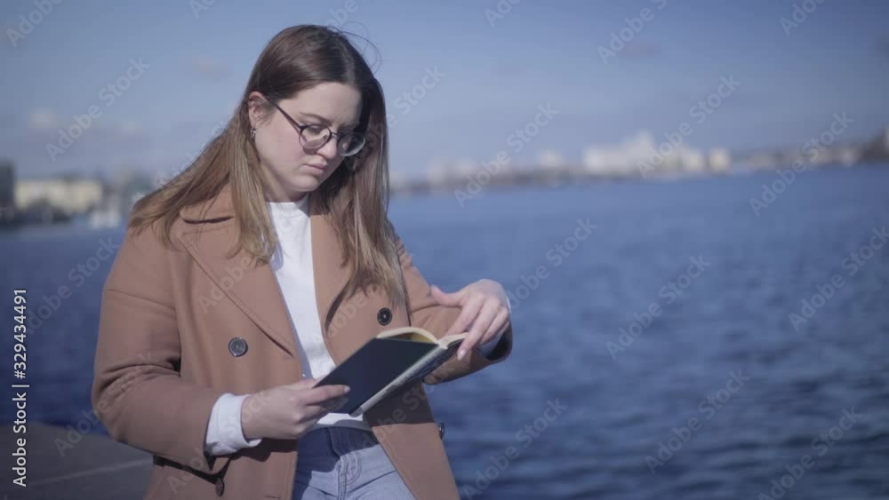 Portrait of young Caucasian woman reading on river bank. Brunette girl in eyeglasses spending autumn day outdoors. Hobby, lifestyle, literature, joy. Portrait of young Caucasian woman reading on river bank. Brunette girl in eyeglasses spending autumn day outdoors. Hobby, lifestyle, literature, joy.