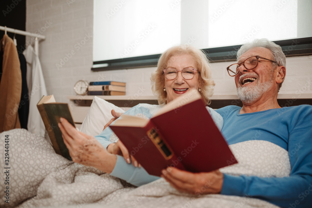 Portrait of good looking elderly couple wearing glasses are reading favorite books while lying in bed at bedroom.