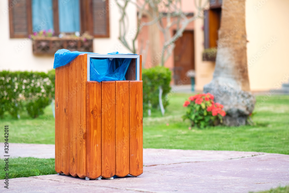 Obraz premium Yellow wooden trash can outdoors on the side of sidewalk in park. Garbage container outside.
