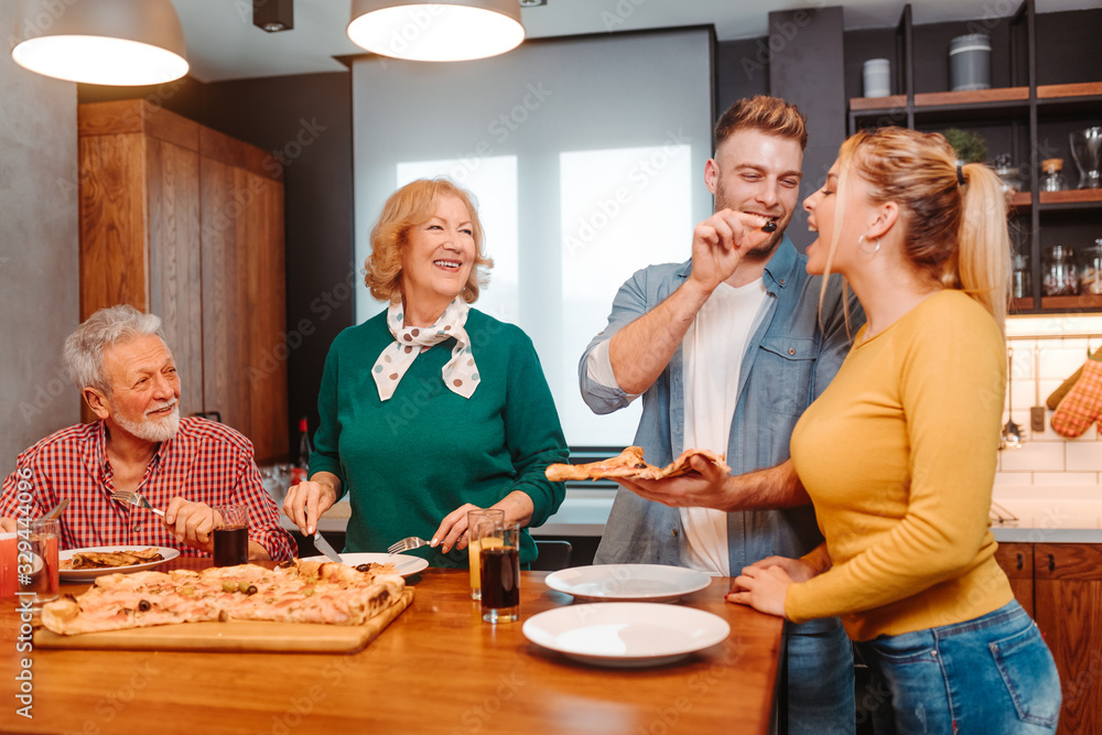 Family is eating pizza in the kitchen. Young man is giving an black olive to his girlfriend. Family is eating pizza in the kitchen. Young man is giving an black olive to his girlfriend.