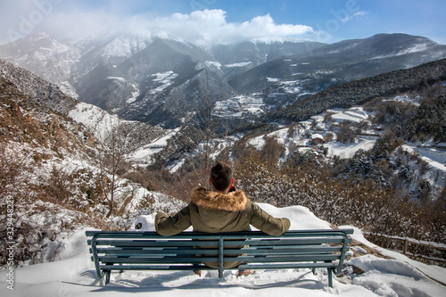Young man sitting on a bench on the side of a mountain enjoying a snowy landscape in the background in winter.