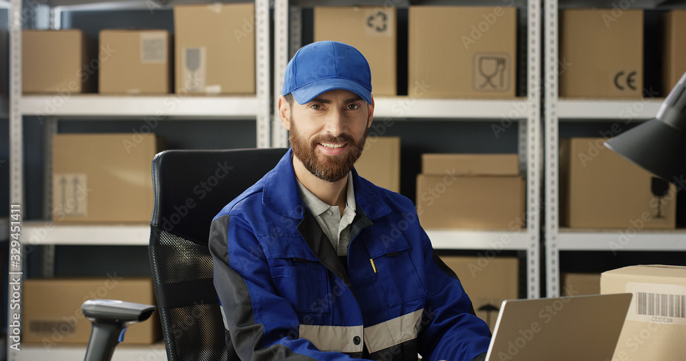 Portrait of young happy Caucasian postman in iniform and cap sitting at desk in postal office store and working at computer. Handsome male courier smiling happily to camera in storage of parcels. Portrait of young happy Caucasian postman in iniform and cap sitting at desk in postal office store and working at computer. Handsome male courier smiling happily to camera in storage of parcels.