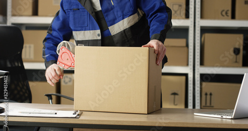 Close up of hands of Caucasian postal male worker in uniform packing carton box at delievery department in post office. Postman closing parcel with sticky tape. Mail shipping concept. photo