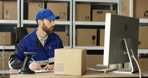 Handsome Caucasian mailman in blue iniform and cap sitting at table in postal office store and working at computer. Male post courier filling in invoice and entering data online in parcel storage. © VAKSMANV