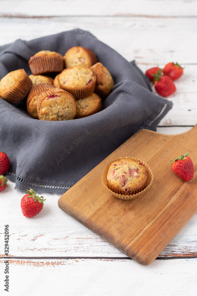 Bowl of Freshly Baked Homemade Strawberry Muffins in a Wood Bowl; One Isolated on Wood Cutting Board in Front; Fresh Strawberrries Scattered Around; White Wood Tabletop