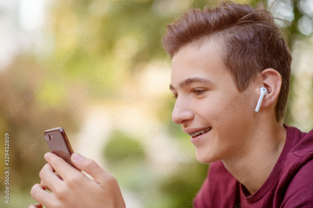 Happy teenage boy is using mobile phone, outdoors.  Close-up portrait of a smiling young man with smartphone, in park.  Cheerful teenager in casual clothes with cell phone in park. Soft focus