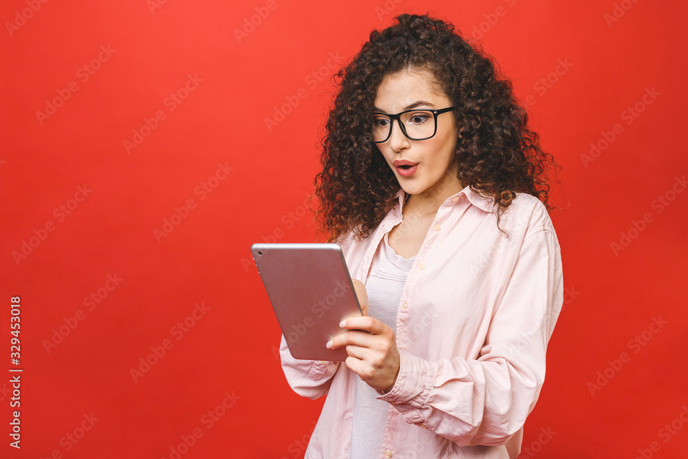 Excited amazed young beautiful woman with tablet pc. Happy girl in white shirt using tablet computer, isolated on red background.