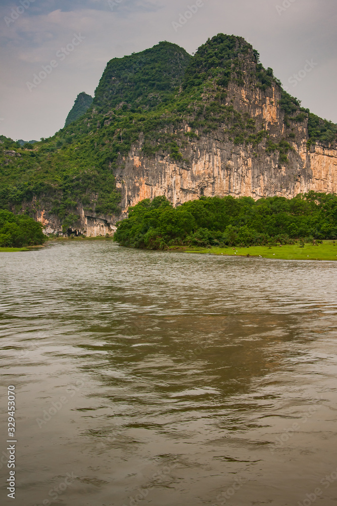 Guilin, China - May 10, 2010: Along Li River. Landscape with green forested karst mountains with brown-black vetical cliff under dark gray sky. Some trees, and Brown water up front.