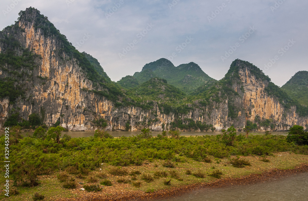 Obraz premium Guilin, China - May 10, 2010: Along Li River. Landscape with green forested karst mountains with brown-black vetical cliffs under blue-white sky, behind land tongue with green bushes.