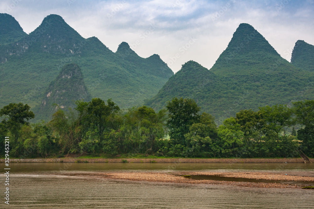 Fototapeta premium Guilin, China - May 10, 2010: Along Li River. Landscape with chain of green forested karst mountain and under blue cloudscape. Green tree belt, Brown water and marsh land up front.