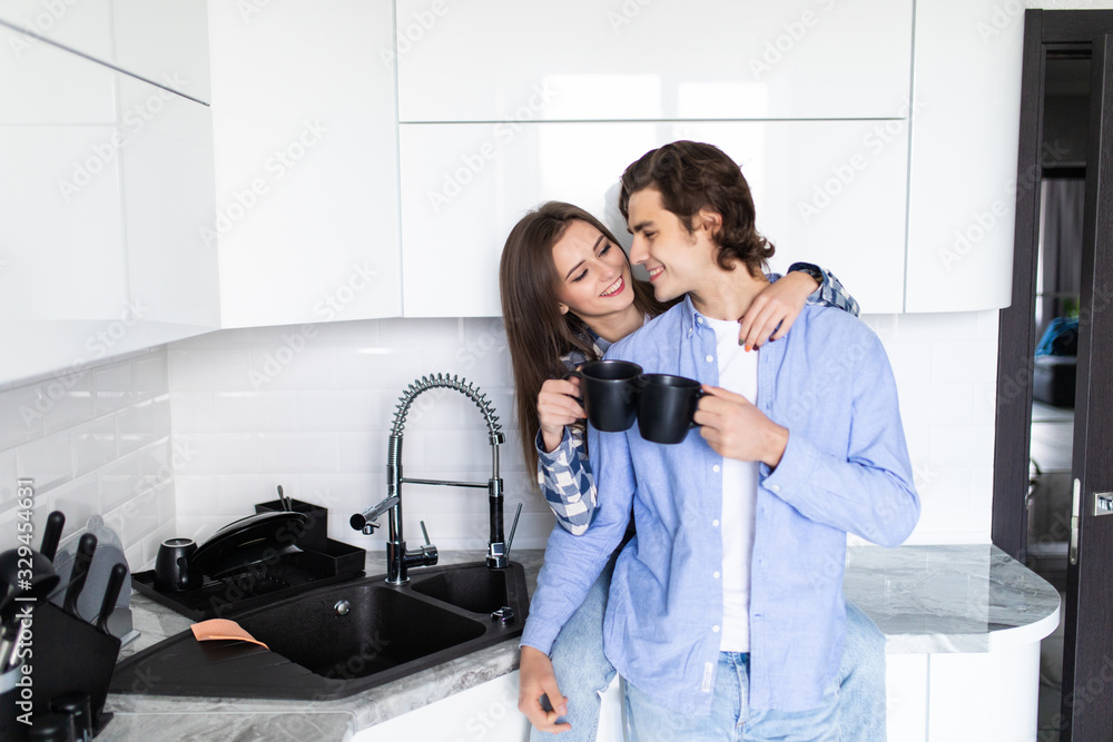 Young man and woman family couple standing in the kitchen together hugging kissing driking coffee holding cups Young man and woman family couple standing in the kitchen together hugging kissing driking coffee holding cups