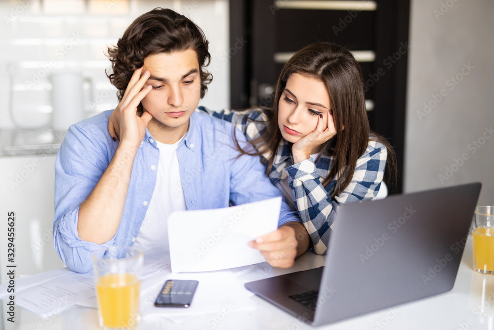 Young married couple with many debts doing paperwork together, reviewing their bills, planning family budget and calculating finances at kitchen table with papers, calculator and laptop computer Young married couple with many debts doing paperwork together, reviewing their bills, planning family budget and calculating finances at kitchen table with papers, calculator and laptop computer