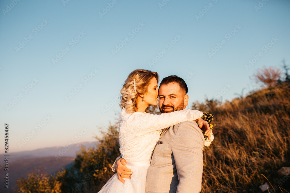 Beautiful couple having a romantic moment on their weeding day, in mountain, sunset.She is in a white wedding dress with a bouquet of sunflowers in hand,groom in a suit. Groom holds bride in his hands Beautiful couple having a romantic moment on their weeding day, in mountain, sunset.She is in a white wedding dress with a bouquet of sunflowers in hand,groom in a suit. Groom holds bride in his hands