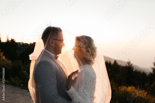 Beautiful couple having a romantic moment on their weeding day, in mountains at sunset. Bride is in a white wedding dress holding bouquet, groom in a suit. Happy hugging couple under the veil.  © Melika