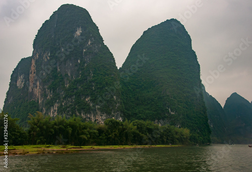 Guilin, China - May 10, 2010: Along Li River. Landscape of rounded twin forested karst mountain peaks under silver sky. Green trees separates them from greenish water. © Klodien