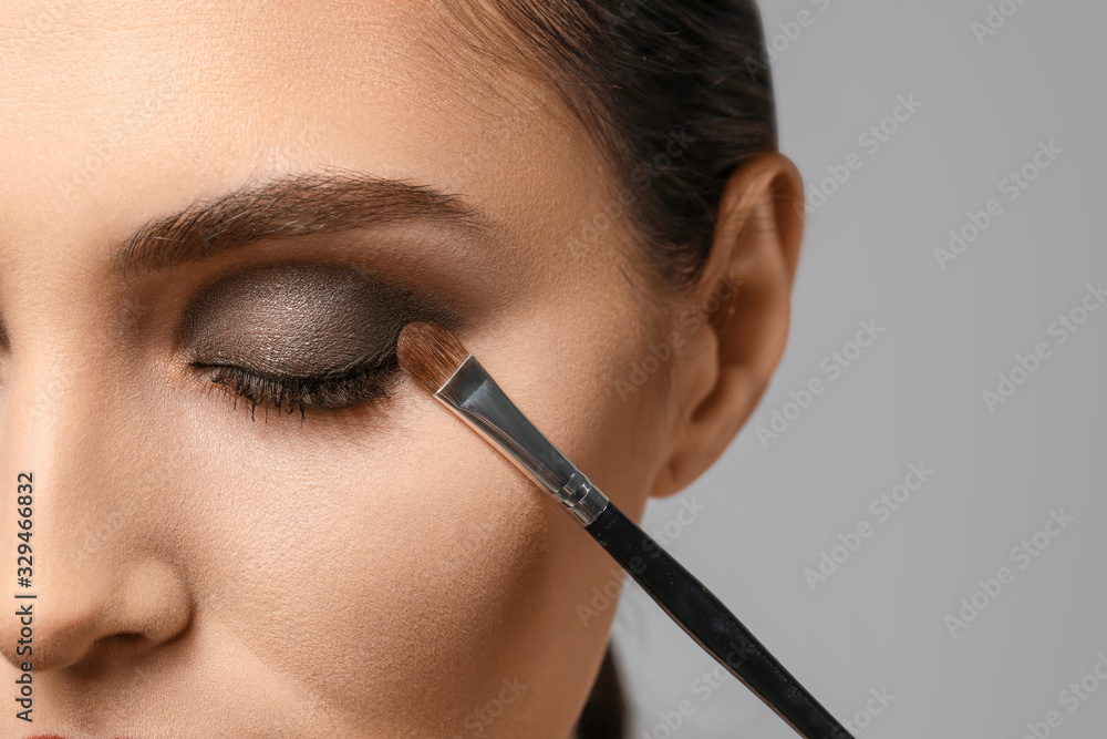 Beautiful young woman applying makeup against grey background, closeup