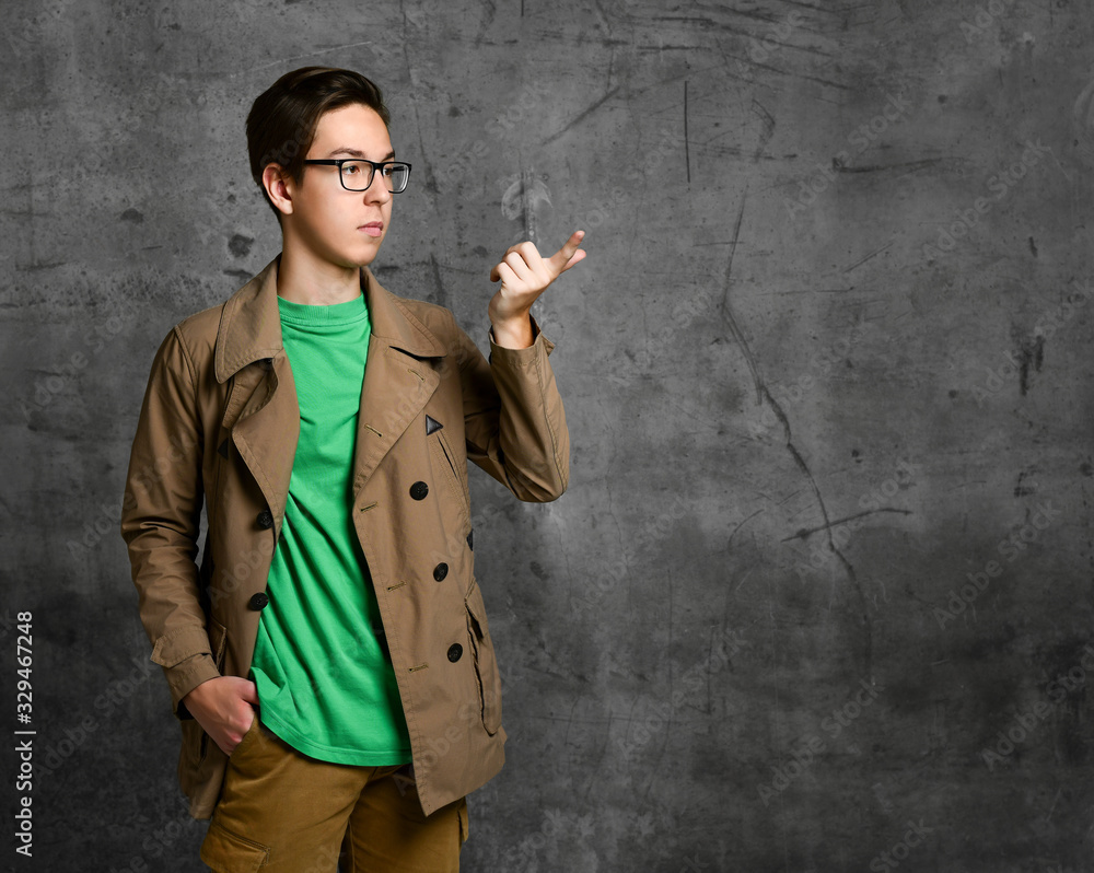 Young teen boy in brown costume and glasses standing and pointing at something with finger over grey concrete wall background Young teen boy in brown costume and glasses standing and pointing at something with finger over grey concrete wall background