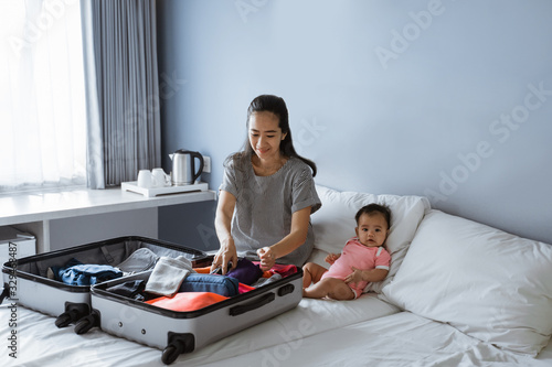 cute baby sits beside the mother preparing clothes to put in the suitcase