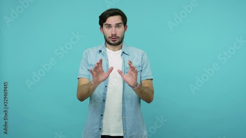 Worried scared panicking man in jeans shirt raising his hands in defensive gesture and looking in horror at camera, walking away from frightening problem. studio shot isolated on blue background