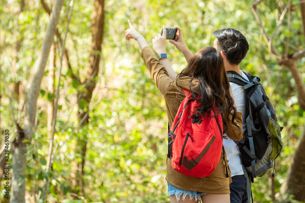 Rear view of happy young Asian couple man and beautiful woman holding hands and hiking together with backpack in forest mountain with using smartphone taking a photo. Outdoors holiday vacation concept