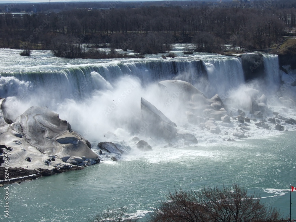 The lovely and lively Niagara Falls with clean, blue water runs down the fall into the river causing a splash over white rocks and boulders and creating a mist in front of the fall with trees behind. The lovely and lively Niagara Falls with clean, blue water runs down the fall into the river causing a splash over white rocks and boulders and creating a mist in front of the fall with trees behind.