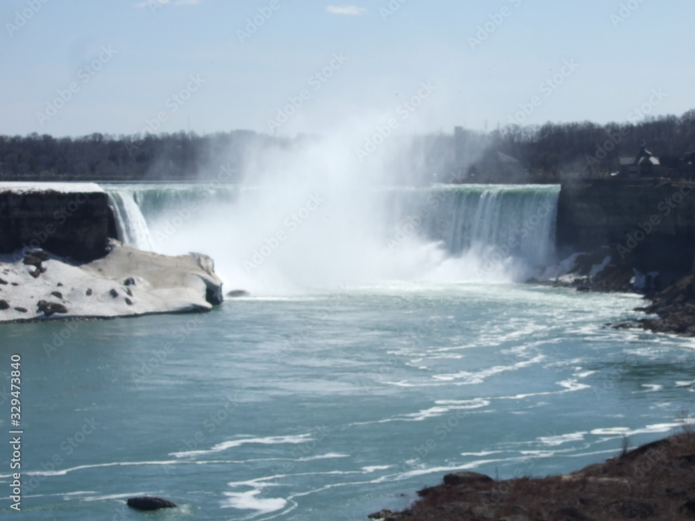 The lovely and lively Niagara Falls with clean, blue water runs down the fall into the river causing a splash over white rocks and boulders and creating a mist in front of the fall with trees behind. The lovely and lively Niagara Falls with clean, blue water runs down the fall into the river causing a splash over white rocks and boulders and creating a mist in front of the fall with trees behind.