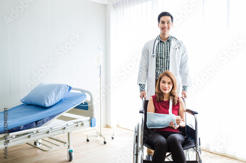 Asian Young man doctor of checking splint the arm of female patient hand due to with her arm broken for better healing with a smile sit in a wheelchair In the room hospital background.