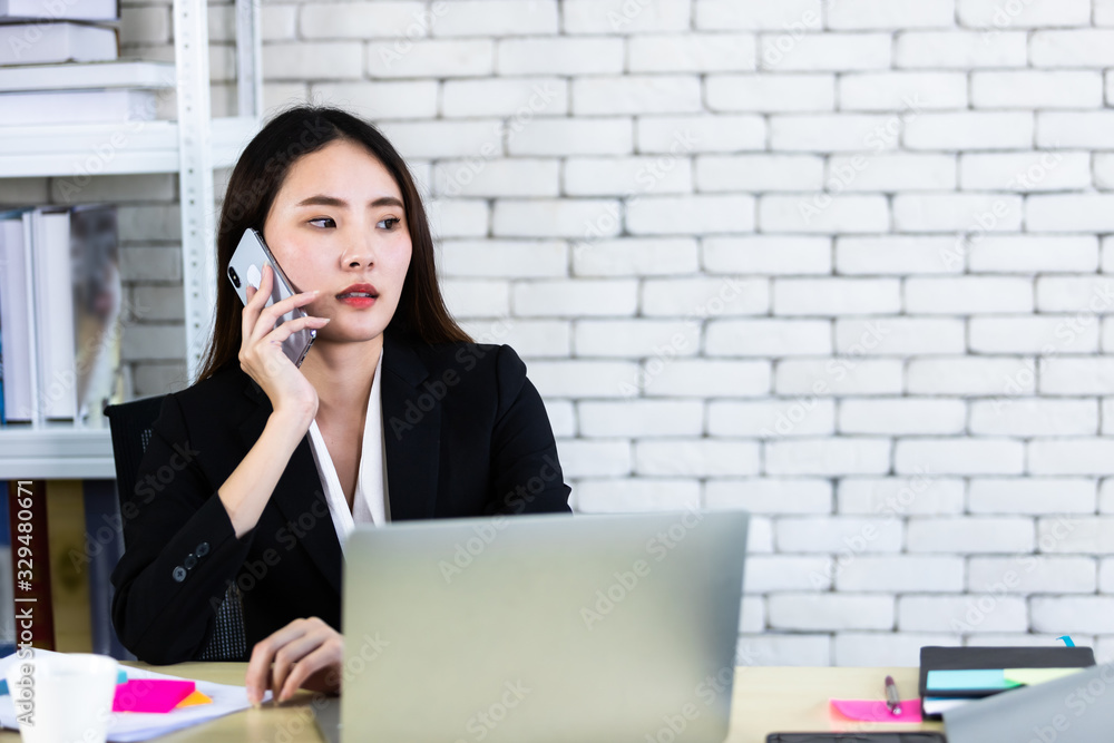 Happy of successful asian young businesswoman hold pick up smartphone working with laptop computer in on White wooden table in white wall office background Happy of successful asian young businesswoman hold pick up smartphone working with laptop computer in on White wooden table in white wall office background