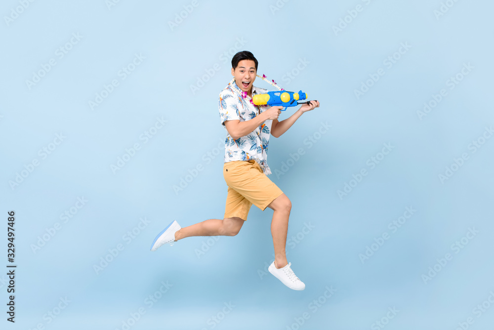 Handsome smiling young Asian man playing with water gun and jumping in studio blue background for Songkran festival in Thailand and southeast Asia Handsome smiling young Asian man playing with water gun and jumping in studio blue background for Songkran festival in Thailand and southeast Asia