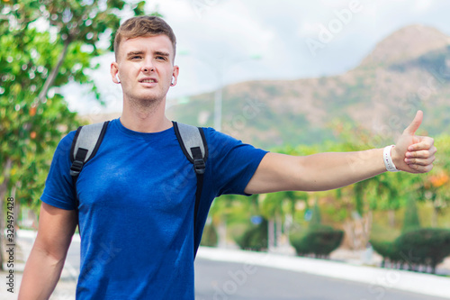 Handsome guy, young man traveler trying to stop, catch taxi car, cab. Boy standing near road with backpack, wireless earphones holding out his hand with thumb up, hitchhiking sign. Hitchhiker, summer
