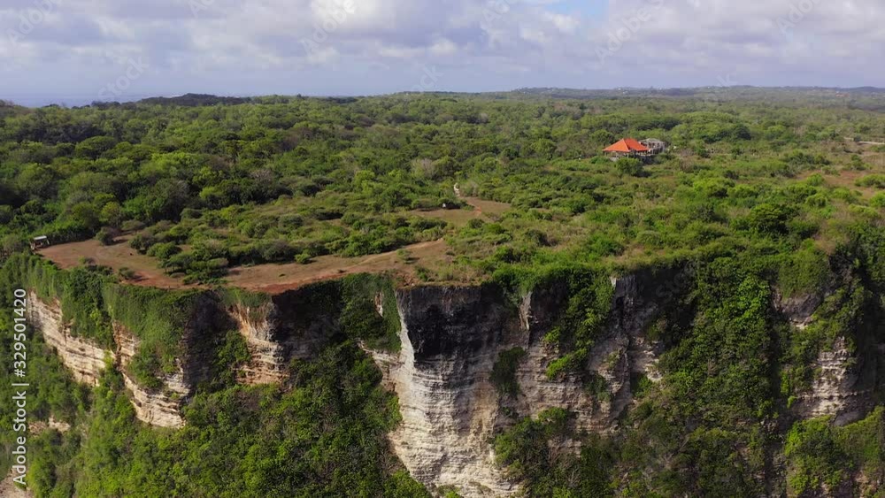 Uluwatu cliffs, revealing the sharp edge Cliff with lush green landscape, tertiary limestone layers and Indian Ocean waves breaking on the beach of Bali, Indonesia. Pull back aerial view. Uluwatu cliffs, revealing the sharp edge Cliff with lush green landscape, tertiary limestone layers and Indian Ocean waves breaking on the beach of Bali, Indonesia. Pull back aerial view.