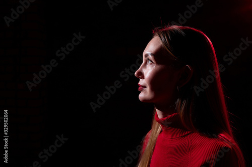 Portrait of elegant woman with long hair in red sweater and dark background with red light. Model posing doring studio photo shot