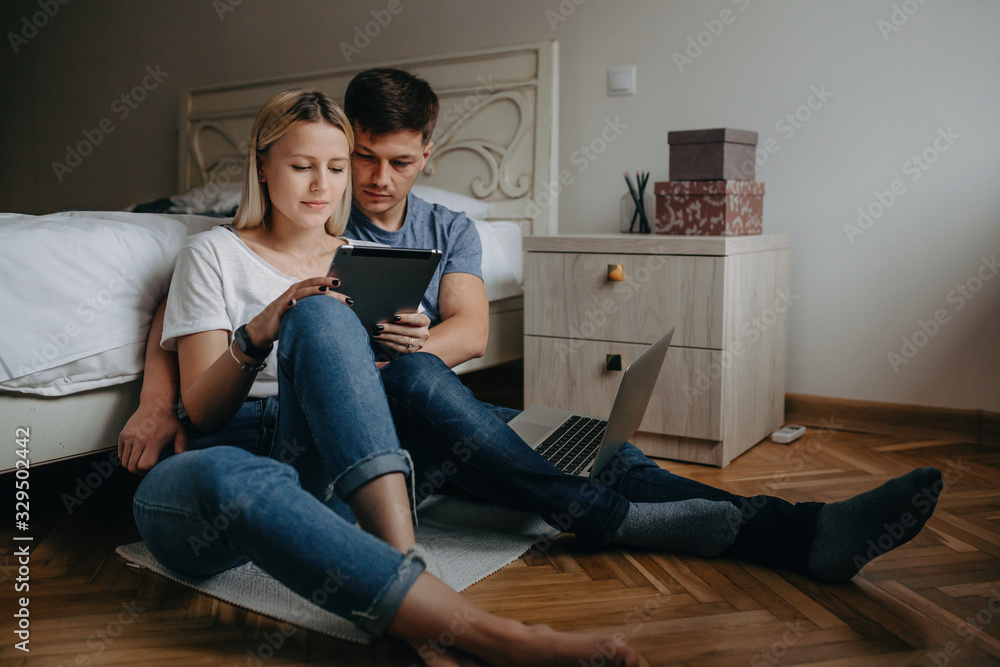 Caucasian couple embracing and browsing on the tablet while sitting on the floor in the bedroom Caucasian couple embracing and browsing on the tablet while sitting on the floor in the bedroom