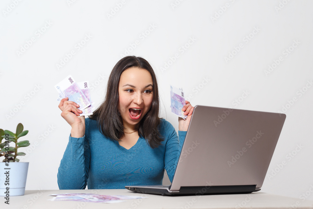 Young cheerful woman holding fan of cash money banknotes, sit and work at desk with pc laptop on white background Young cheerful woman holding fan of cash money banknotes, sit and work at desk with pc laptop on white background