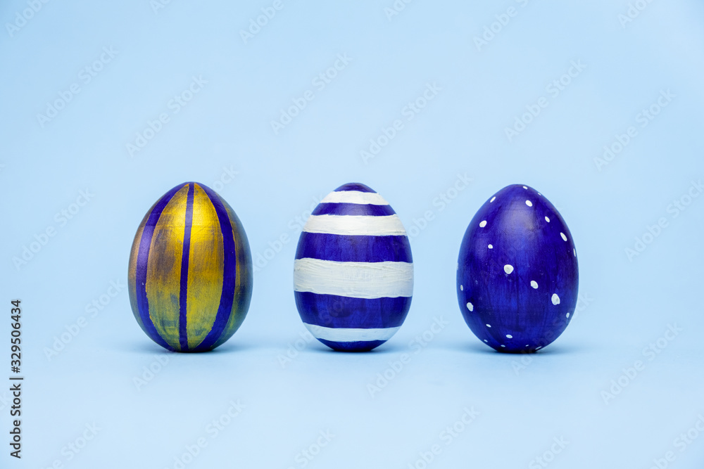 Fototapeta premium Three easter trendy colored classic blue and golden decorated eggs striped pattern on blue. Happy Easter card with copy space for text. Minimal style.
