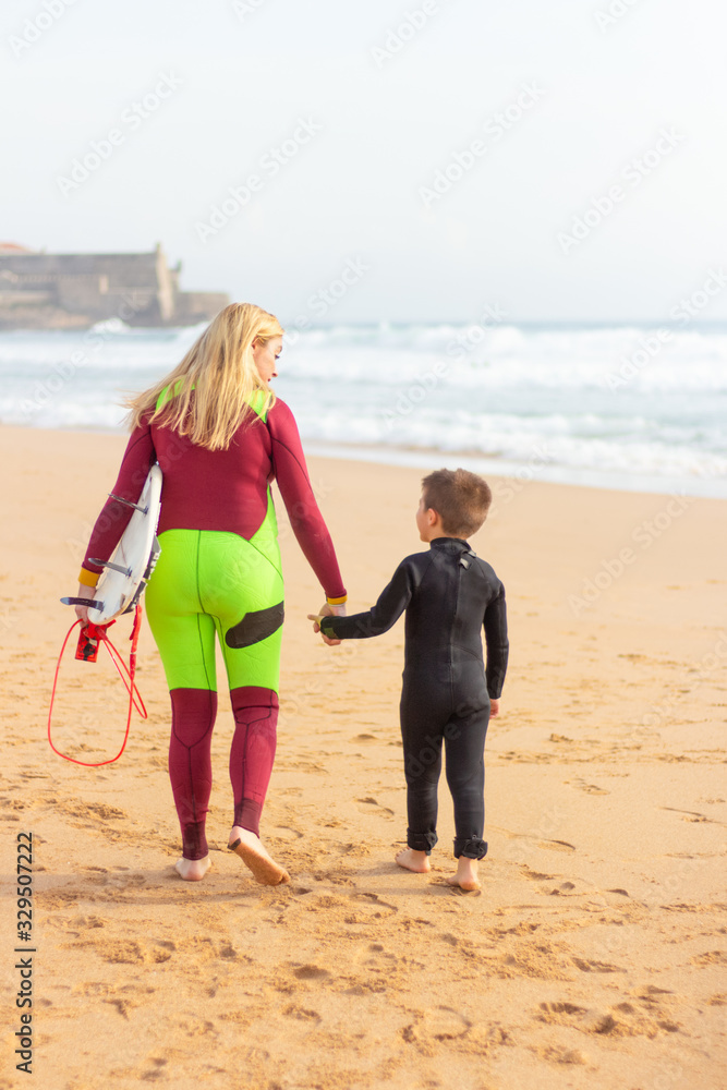 Back view of mother and son with surfboard on beach. Rear view of mother and cute little son in wetsuits walking together on sandy beach. Surfing concept Back view of mother and son with surfboard on beach. Rear view of mother and cute little son in wetsuits walking together on sandy beach. Surfing concept