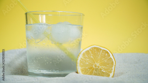 glass with ice and soda water and lemon on the sand on a yellow background
