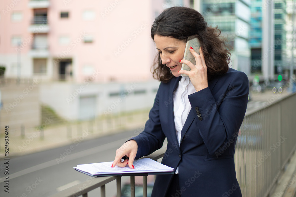 Businesswoman talking by smartphone and looking at papers. Focused middle aged businesswoman talking via cell phone and looking at folder with documents on street. Business concept Businesswoman talking by smartphone and looking at papers. Focused middle aged businesswoman talking via cell phone and looking at folder with documents on street. Business concept