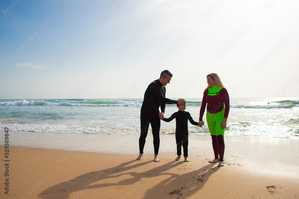 Family holding hands on sandy beach. Cheerful parents in wetsuits looking at adorable smiling little son on ocean coast. Family time concept