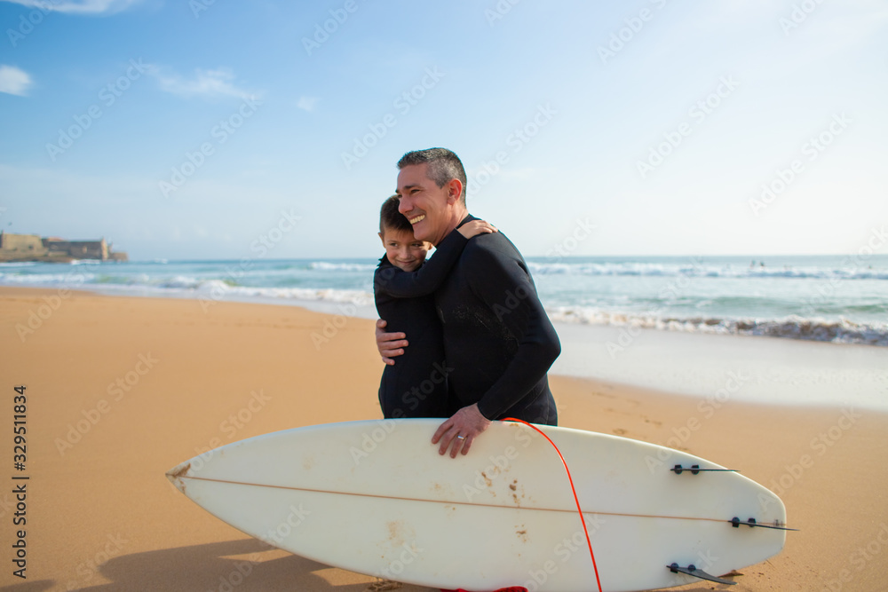 Obraz premium Father and son in wetsuits hugging on beach. Cheerful father and son hugging while spending time together with surfboard on beach. Water sport concept