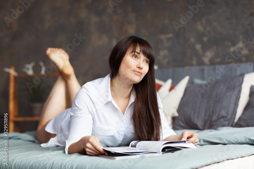 Young beautiful happy woman reading a magazine in the bedroom. Portrait of a girl in a white shirt lying on the bed with a book in her hands © Sviatlana
