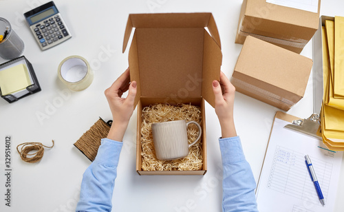 delivery, mail service, people and shipment concept - female hands packing mug to parcel box with straw filler at post office photo