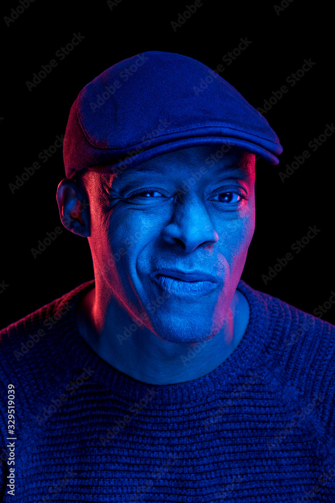 Black man with blue and red light, wearing a flat cap, isolated on black background, looking at the camera. Uncertainty expression. Vertical