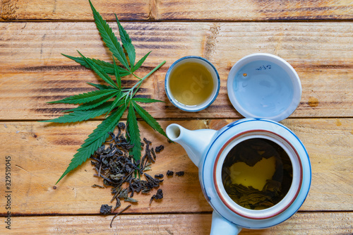 The white and blue teapot, a tea cup, green marijuana leaves and dried marijuana leaves placed on an old wooden table. top view.
