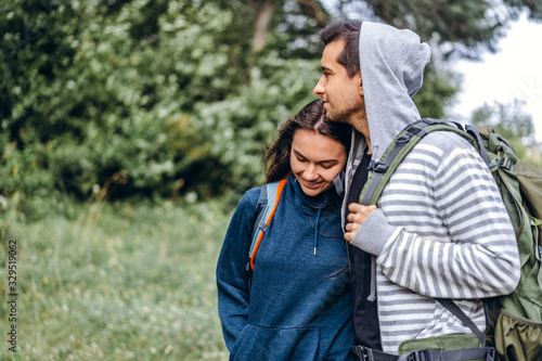 Young couple with backpacks on their backs in the forest. Loving man kisses his beautiful girlfriend on a hike in the woods
