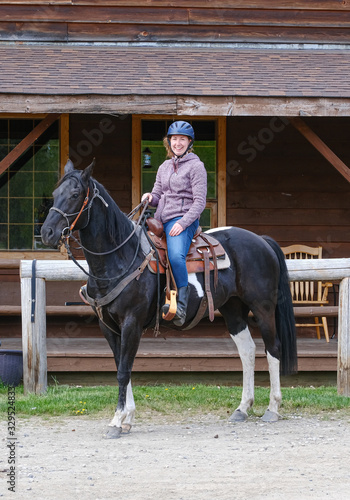 A young woman riding a black and white horse in front of a wooden house, Banff national park © Dasya - Dasya